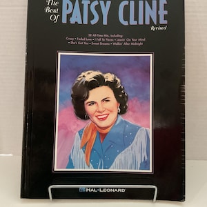 May include: A black music book titled "The Best of Patsy Cline" with a portrait of Patsy Cline. The book includes the text "Revised" and lists 38 all-time hits, including "Crazy" and "Faded Love". The book is held by a metal stand.