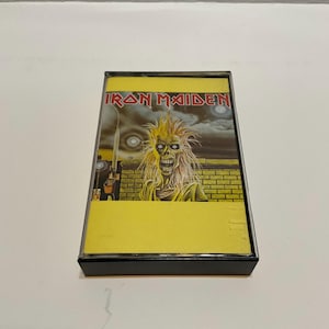 May include: A vintage cassette tape featuring the album art for Iron Maiden. The artwork depicts a skeletal figure with yellow hair and a menacing expression against a brick wall and cloudy sky. The cassette has a yellow label.