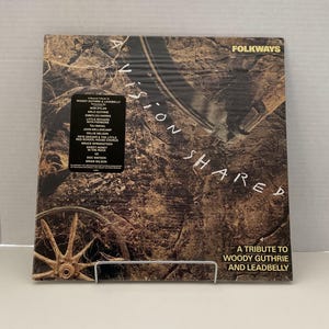 May include: A vinyl record album titled "A Vision Shared" with the Folkways logo. The album cover features a sepia-toned image with the text "A Tribute to Woody Guthrie and Leadbelly." The album includes artists such as Bob Dylan and Bruce Springsteen.