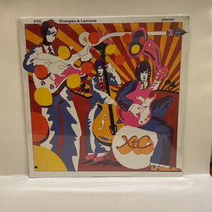May include: A vinyl record album titled "Oranges & Lemons" by XTC. The album cover features a colorful illustration of a band playing instruments, with a retro, psychedelic design in shades of red, orange, yellow, pink, and blue.