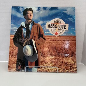 Peut inclure: Pochette d'album pour "Absolute Torch and Twang" de k.d. lang and the Reclines. L'illustration montre une personne en manteau marron et chapeau, debout dans un champ sous un ciel bleu. Le titre de l'album est dans une bannière.