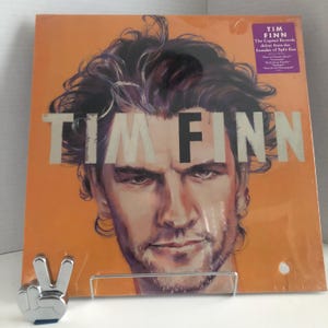Peut inclure: Une pochette d'album vinyle avec un portrait de Tim Finn, le fondateur de Split Enz. La pochette est orange avec une image en noir et blanc de Tim Finn. Le titre de l'album "Tim Finn" est imprimé en grandes lettres blanches en haut de l'image.