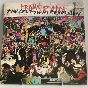 May include: A vintage vinyl record album cover featuring a colourful, chaotic scene of people in a crowded room. The album title "Frank Zappa Tinseltown Rebellion" is written in bold, white letters across the top of the cover.