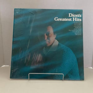 May include: A vintage vinyl record album titled "Dion's Greatest Hits." The album cover features a portrait of Dion, with the album title in white text. The background is a teal blue color.