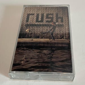 May include: A vintage cassette tape featuring the album cover for "Roll the Bones" by Rush. The artwork depicts a person kicking a skull, with the band's name in dice-shaped letters. The background shows a wall of dice and water.