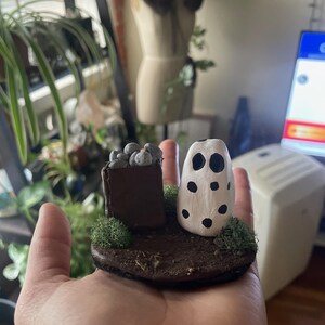 May include: A miniature Halloween scene with a white ghost with black dots, a brown box with black berries, and green moss on a brown base.