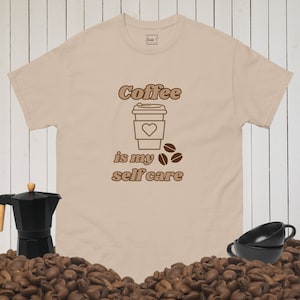 Può includere: Maglietta beige con testo marrone che recita "Coffee is my self care". Il testo è in un font retrò. Un contorno marrone di una tazza di caffè con un cuore sopra è sopra il testo. Tre chicchi di caffè marroni sono a destra del testo.