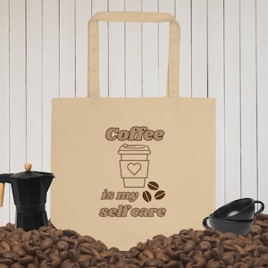 May include: A beige tote bag featuring a brown outline of a coffee cup with a heart inside and the text "Coffee is my self care" printed on it. The bag is surrounded by coffee beans.