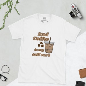 Può includere: Maglietta bianca con una grafica marrone di un bicchiere di caffè freddo con una cannuccia e il testo "Iced Coffee is my self care".