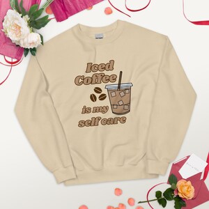 May include: A beige sweatshirt with the text "Iced Coffee is my self care" in brown lettering. A cartoon drawing of a glass of iced coffee with a straw and ice cubes is next to the text.