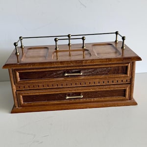 May include: A rectangular wooden chest with two drawers, each featuring a thin horizontal brass handle. The top surface has three recessed compartments and a decorative brass railing along the back edge. The wood has a medium brown finish.