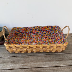 May include: Rectangular woven basket with rope handles. The interior is lined with a floral fabric featuring a colorful pattern of small flowers on a brown background. The basket sits on a wooden surface.