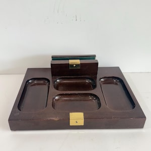 May include: A dark brown wooden desk organizer featuring three rectangular recessed compartments and a central upright section with a green interior lining. The piece is accented with two polished gold-tone metal hardware plates.
