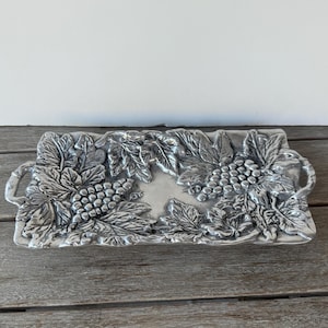 May include: A rectangular silver-colored serving tray with handles, adorned with detailed grape and leaf designs. The tray's edges are slightly raised, and the grape clusters and leaves are intricately sculpted, creating a textured surface. The tray is approximately 12 inches long.