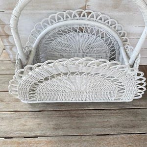 May include: A white wicker basket with a curved handle and scalloped edges. The basket has a round, woven interior and is set against a weathered wooden background. The basket is empty.