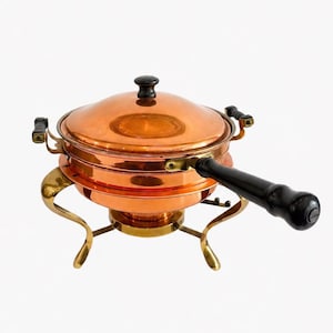 May include: A copper fondue pot with a black handle and lid knob. The pot sits on a brass stand with curved legs. The pot has two small handles on either side.