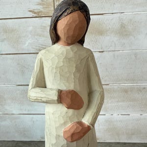 May include: A carved wooden figurine of a person in a long, cream-colored dress. The figure has a brown head covering and arms, with hands clasped over the abdomen. The figurine has a textured surface.