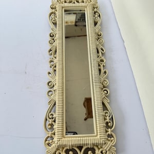 May include: A long, rectangular mirror with an ornate, cream-colored frame. The frame features intricate scrollwork and a ribbed inner border. The mirror reflects a room's interior. A decorative accent for any space.