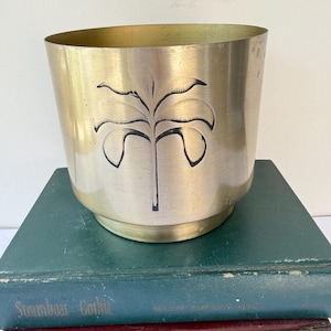 May include: A brass-colored metal planter with a black palm tree design. The planter sits on a stack of vintage books with green and red covers. The planter has a cylindrical shape with a slightly flared top.