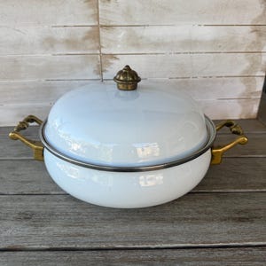 May include: White enamelware pot with a lid and brass-colored handles. The pot has a rounded shape and a brass knob on the lid. The pot is sitting on a wooden surface.