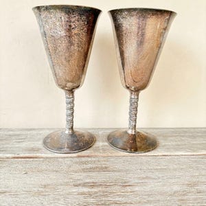 May include: Two vintage silver goblets with a textured, aged appearance. The goblets have a flared bowl shape and a twisted stem design. The bases are round and slightly worn, suggesting age and use. The goblets are displayed on a weathered wooden surface.