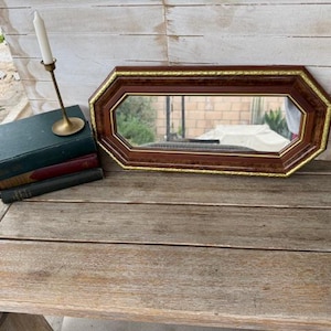 May include: A rectangular, brown framed mirror with a gold trim sits on a wooden table. A stack of three books and a brass candlestick with a white candle are next to the mirror. The mirror reflects a view of a yard.