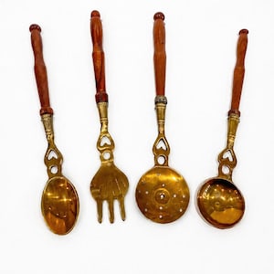 May include: Four vintage brass kitchen utensils with wooden handles. The set includes a spoon, fork, slotted spoon, and ladle. The brass has a warm, golden hue, and the wooden handles are a rich brown color.