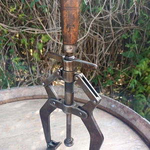 May include: A vintage three-jaw gear puller featuring a weathered wooden handle, a central threaded metal rod, and three adjustable steel arms with pivoting feet. The mechanical tool displays a dark, oxidised metal finish and a worn wooden grip.