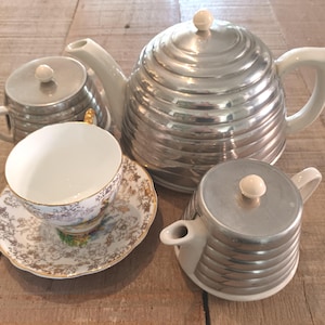 May include: A tea set comprising a large beehive-shaped teapot with a silver-toned ribbed body and white handle, a matching lidded sugar bowl, a lidded creamer, and a single teacup and saucer featuring a delicate floral pattern with gold trim.