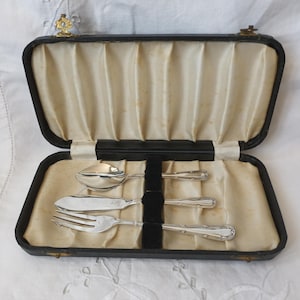 May include: A set of three silver-toned utensils including a spoon, a knife, and a fork, arranged horizontally inside a rectangular black case. The interior of the case is lined with cream-coloured pleated fabric and features a black velvet divider.