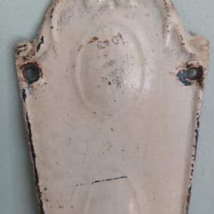 May include: An antique, cream-coloured metal door plate with an ornate, arched top. The plate has two holes for mounting and shows signs of wear, including chipped paint and rust. The design features a central oval shape and decorative flourishes.