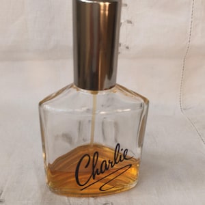 May include: A clear glass perfume bottle with a silver cap and the word "Charlie" in black script. The bottle contains a golden-coloured liquid. The bottle has a geometric shape.