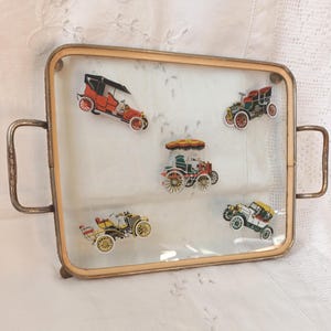 May include: A rectangular glass serving tray with a metallic frame and two side handles. The tray features five colourful illustrations of vintage automobiles, including red, green, yellow, and white models, arranged across the clear glass surface.