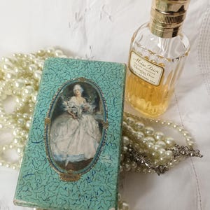 May include: A vintage perfume bottle and a small, rectangular box with a portrait of a woman in a white dress. The box is teal with a repeating pattern. A strand of faux pearls is draped around the items.