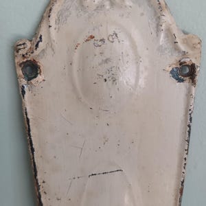 May include: An antique, cream-coloured metal door plate with an ornate design. The plate shows signs of wear and tear, including rust and chipped paint. There are two holes for mounting. The overall effect is one of age and use.