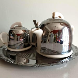May include: A tea set consisting of a teapot, a sugar bowl, and a creamer, all featuring a combination of cream-coloured ceramic bases and polished chrome metal covers, arranged together on a round, decorative silver-toned metal serving tray.