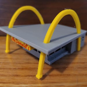May include: A gray and yellow plastic model of a McDonald's restaurant with the iconic golden arches. The model has a drive-thru window and the words "McDonald's" printed on the front.