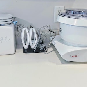 Puede incluir: Un tarro de cerámica blanco con la palabra "thankful" escrita en él, una batidora de pie blanca con un tazón plateado y una perilla plateada, y un soporte de accesorios de batidora de pie blanco con tres accesorios.