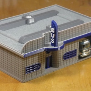 Peut inclure: Une gare routière miniature en gris et bleu avec un toit gris et une enseigne bleue indiquant "BUS". Un petit bus miniature est garé à l'intérieur de la gare.