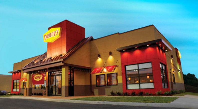 May include: Exterior view of a Denny's restaurant at dusk. The building is yellow with red accents and a large sign with the Denny's logo in red and yellow.