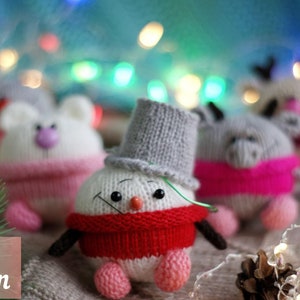 May include: A knitted snowman toy with a red scarf and a grey hat. The snowman is sitting on a light brown surface with other knitted toys in the background.
