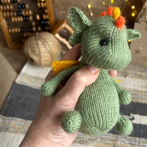 Può includere: Un giocattolo di drago di lana verde con accenti arancioni e gialli. Il drago ha un corpo piccolo e rotondo e una lunga coda. Viene tenuto nella mano di una persona.
