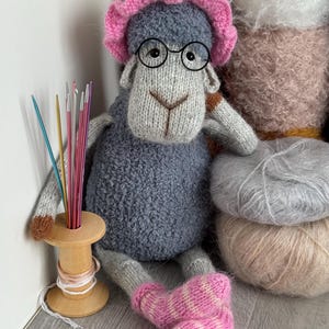 Puede incluir: Muñeca de oveja tejida a mano, cuerpo gris, gorro rosa y gafas. La oveja está sentada junto a una bobina de madera con agujas de tejer e hilo. Hay ovillos de lana en el fondo. La muñeca lleva calcetines a rayas rosas y blancas.