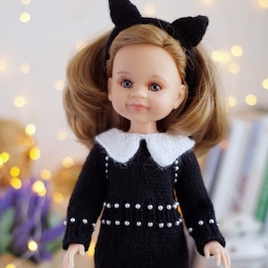 Peut inclure: Une poupée portant une robe noire en tricot avec un col blanc et des accents de perles, un bandeau d'oreilles de chat noir et des chaussures blanches. La poupée a les cheveux bruns et les yeux bleus.