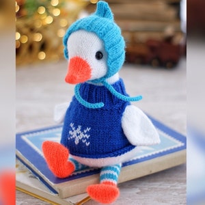 May include: A white knitted goose wearing a blue knitted hat and sweater with a white snowflake design. The goose has orange feet and is sitting on a stack of books.