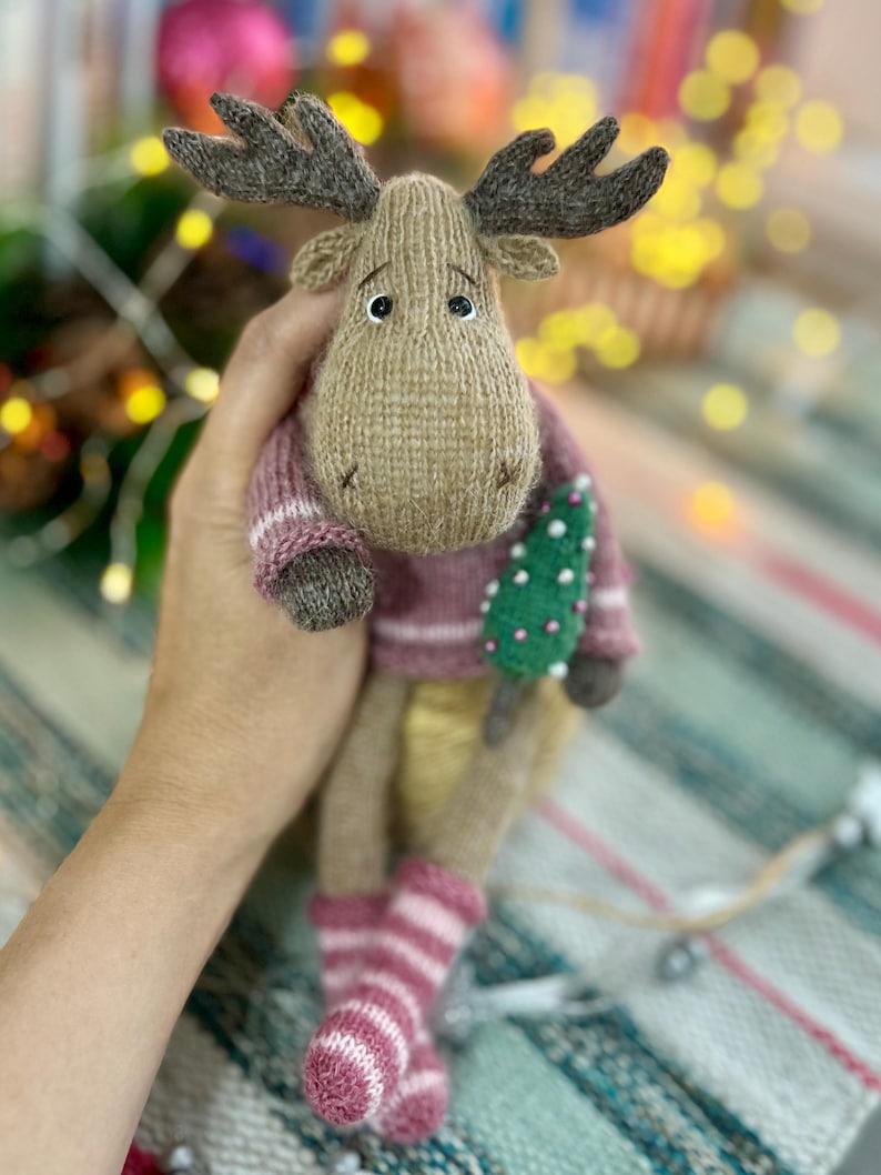 May include: A brown knitted moose toy wearing a pink and white striped sweater and socks, holding a small green Christmas tree.