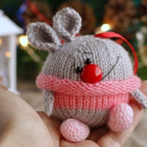 Könnte beinhalten: Eine runde, handgestrickte graue Tierfigur als Ornament mit zwei aufrechten Ohren, einer leuchtend roten Knopf-Nase und einem aufgestickten schwarzen Lächeln. Sie trägt einen rosa gerippten Pullover um die Mitte und hat zwei kleine, runde, hellrosa Füße.