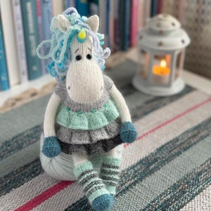 May include: A handmade, knitted unicorn doll with a white body, blue and turquoise mane, and a yellow horn. The unicorn wears a gray, mint green, and black layered dress and striped leg warmers. It sits on a ball of yarn, with a lantern in the background.