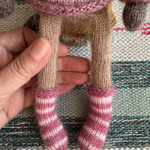 May include: A knitted doll with pink and white striped socks. The doll is wearing a pink sweater and has brown yarn hair.