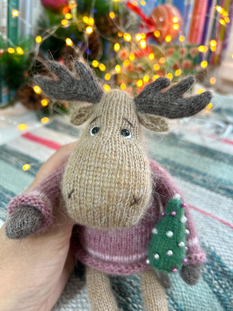 May include: A knitted moose toy wearing a pink sweater and holding a green Christmas tree with white beads. The moose has brown antlers and a brown nose.